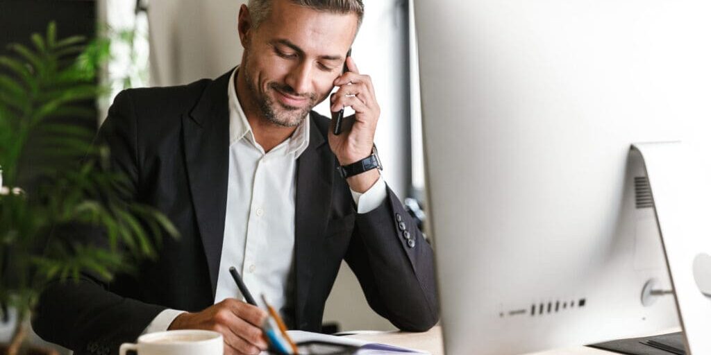 Image of handsome businessman 30s wearing suit talking on cell phone while working on computer in office