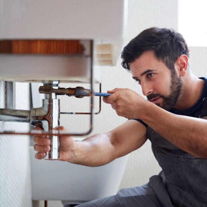 Male Plumber Using Wrench To Fix Leaking Sink In Home Bathroom